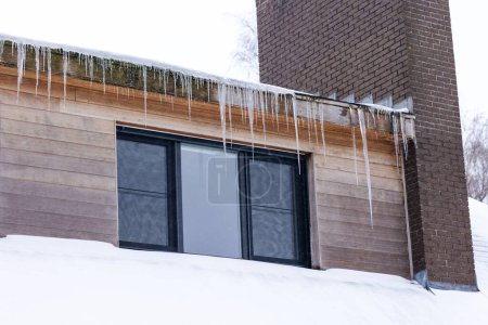 Foto de Un retrato de una casa con enormes carámbanos colgando de la cuneta del techo por encima de una sola ventana junto a una chimenea de un techo, el techo está cubierto de nieve blanca durante el invierno después de una ventisca. - Imagen libre de derechos
