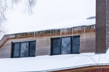 Téléchargez les photos : Portrait d'une maison avec de grandes glaçons suspendus à la gouttière du toit au-dessus de deux fenêtres à côté d'une cheminée d'un toit, le toit est recouvert de neige blanche pendant l'hiver après un blizzard. - en image libre de droit