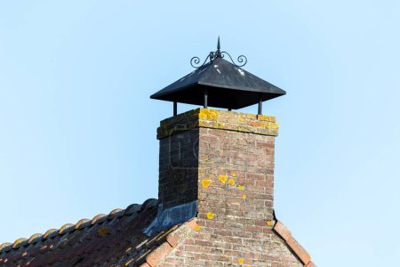 Téléchargez les photos : Portrait d'une cheminée en brique grise surmontée d'un petit toit afin que la pluie n'y tombe pas sur un toit carrelé rouge au ciel bleu. - en image libre de droit