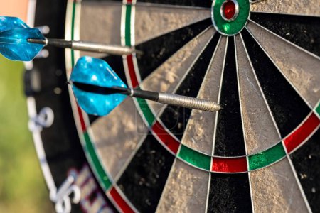 A closeup portrait of a part of a dartboard game standing outdoors on a sunny day, with two darts sticking in it. the bull is also visible. the darts are ready to be thrown in a game or match.