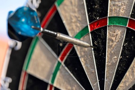 A portrait of a part of a dartboard game standing outdoors on a sunny day, with one dart sticking in it. the bull is also visible. the darts are ready to be thrown in a game or match.