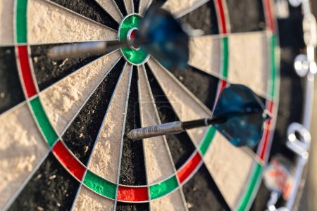 A portrait of a part of a dartboard game standing outdoors on a sunny day, with two darts sticking in it. the bull is also visible. the darts are ready to be thrown in a game or match.
