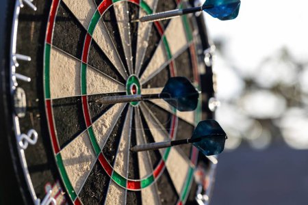 A portrait of a dartboard game outside on a sunny day, with darts sticking in it. the bull is also visible. the darts are ready to be thrown in a game or match.
