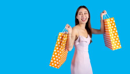 Foto de Happy smiling Asian female shopper holding a bunch of shopping bags while walking and looking at camera. Fashionable girl in dress isolated on blue color background. Half body length in studio shot. - Imagen libre de derechos