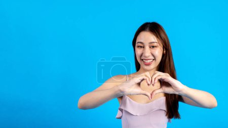 Foto de Happy smiling Asian female stylish woman making a heart hand gesture isolated in blue background. Expressive girl showing love in half body length studio shot - copy space - Imagen libre de derechos