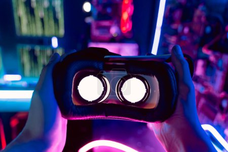 Téléchargez les photos : Eyes perspective of person hands holding 3d 360 virtual reality headset wear, augmented reality goggles in neon light futuristic environment. VR AR innovation technology background - en image libre de droit