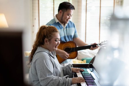 Photo for Young Asian chubby couple singing and playing acoustic guitar and piano together. Man and woman enjoying musical instrument. People in a band practicing in the house - Royalty Free Image