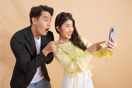 Téléchargez les photos : Shocked excited Asian businessman along with happy smiling woman pointing finger at smart phone over beige studio background. Man in white shirt and suit and woman in yellow top and white trousers. - en image libre de droit