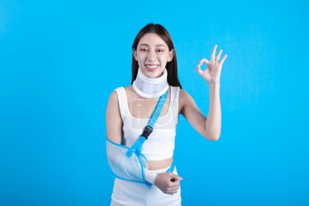 Téléchargez les photos : Beautiful young asian woman with broken arm in soft splint suffering a sore arm showing okay sign isolated on blue background, accident insurance concept. - en image libre de droit