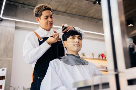 Photo for Happy young Asian male hairdresser smiling wearing apron in modern salon holding brush and scissors trimming hair of client - Royalty Free Image