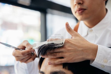 Téléchargez les photos : Jeune et beau salon de coiffure homme taille les cheveux en utilisant peigne et ciseaux tout en se concentrant sur le travail dans le salon moderne - en image libre de droit