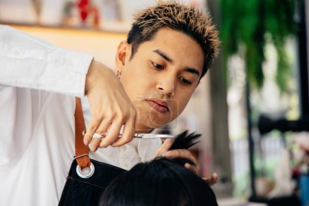 Foto de Joven y guapo asiático peluquero masculino recorte mans cabello usando peine y tijeras mientras se concentra en el trabajo en el salón moderno - Imagen libre de derechos