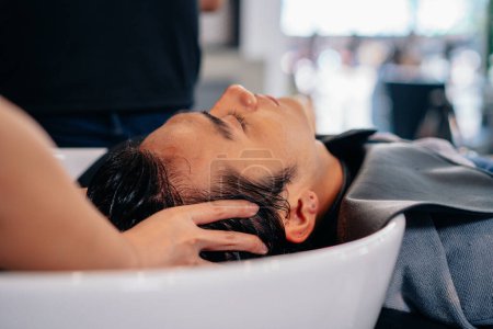 Téléchargez les photos : Beau jeune homme asiatique relaxant avec les yeux fermés avec la tête dans l'évier tandis que les mains de coiffeuse rinçant les cheveux et pressant les points de pression - en image libre de droit