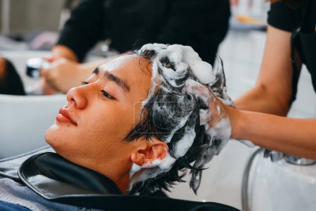 Téléchargez les photos : Beau jeune homme asiatique relaxant avec les yeux fermés avec la tête dans l'évier tandis que les mains de coiffeuse rinçant les cheveux et pressant les points de pression - en image libre de droit