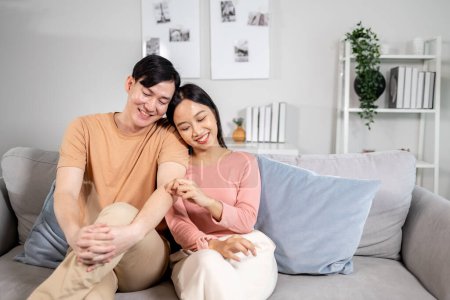 Téléchargez les photos : Jeune asiatique marié heureux couple souriant et assis sur le canapé dans le salon à la maison. Petit ami et petite amie embrassant et relaxant - en image libre de droit