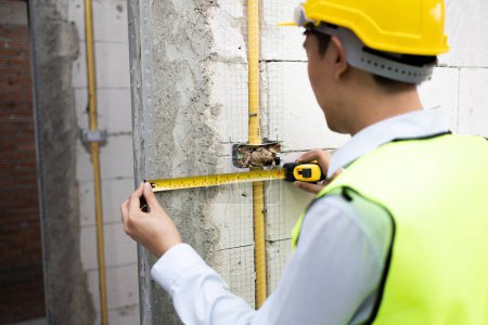 Photo for Young Asian engineer in engineering uniform and helmet at construction site using a measurement tape to measure for precision. 30s professional foreman working on structure building at workplace - Royalty Free Image