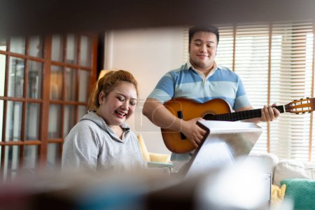 Téléchargez les photos : Young Asian chubby couple singing and playing acoustic guitar and piano together. Man and woman enjoying musical instrument. People in a band practicing in the house - en image libre de droit