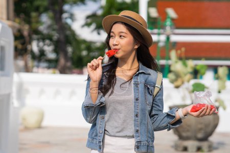 Photo for Portrait of Asian female traveler eating a slice of water melon while walking on sidewalk of buddhist temple on street in Bangkok, Thailand, Southeast Asia - Woman enjoying street food lifestyle - Royalty Free Image