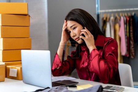 Photo for Young Asian business female entrepreneur having a phone call problem with customer on the phone while using the computer laptop with a stack of packages. Business woman doing online fashion store. - Royalty Free Image