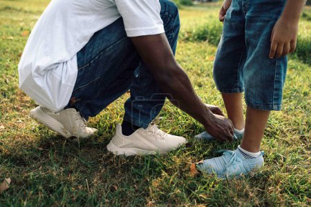 Foto de Día cálido y soleado en el parque: el padre afroamericano ayuda a su hijo a atar cordones de zapatos - Imagen libre de derechos