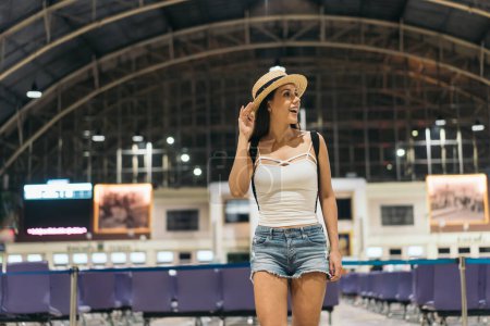 Téléchargez les photos : Vue de face de la jeune voyageuse debout avec sac à dos et chapeau à la gare à la recherche d'un panneau pour se rendre à destination pendant les vacances. - en image libre de droit