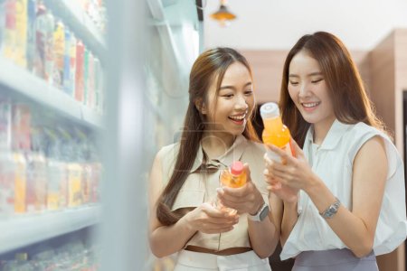 Photo for Two beautiful Asian women shopping and checking items in supermarket. Female friends meeting and examining nutrition labels at convenient store - Royalty Free Image