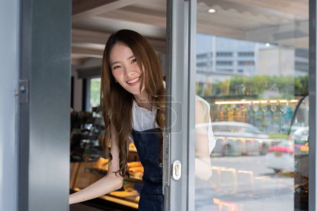 Foto de Joven barista asiática adulta dando la bienvenida a un cliente que entra en la cafetería con una sonrisa amistosa y una cálida bienvenida. Dueño de una pequeña empresa en Asia. El signo de idioma tailandés en la puerta es deslizarse - Imagen libre de derechos