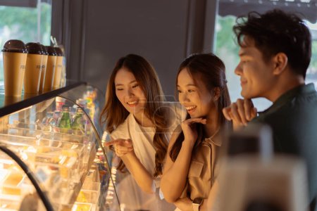 Foto de Jóvenes mujeres asiáticas mirando el pan y los postres mientras están de pie desde la pantalla de la ventana de la panadería con amigos masculinos y femeninos. Grupo de adolescentes de Asia señalando y ordenando en café cafetería - Imagen libre de derechos