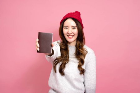 Téléchargez les photos : Belle asiatique femme passager en pull, jeans et bonnet chapeau. Portrait d'une jeune fille souriante montrant un passeport de demi-longueur. Style de vie hivernal et concept de voyage - en image libre de droit