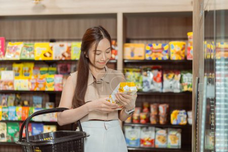 Photo for Young beautiful Asian woman shopping and choosing items in supermarket. Female white collar worker picking something to drink at convenient store - Royalty Free Image
