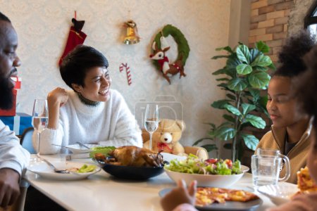 Téléchargez les photos : Portrait de jeune femme avec mari afro-américain appréciant le déjeuner de Noël de dinde à la maison décorée avec des bas, canne à bonbons, couronne, cadeaux et cloches - en image libre de droit