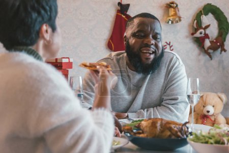 Photo for Portrait of young man with african american husband enjoying christmas lunch of turkey at decorated home with stockings, candy cane, wreath, gifts and bells - Royalty Free Image
