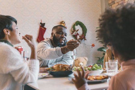 Téléchargez les photos : Portrait de jeune homme avec mari afro-américain profitant du déjeuner de Noël de dinde à la maison décorée avec des bas, canne à bonbons, couronne, cadeaux et cloches - en image libre de droit