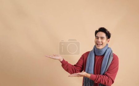 Téléchargez les photos : Heureux et positif asiatique l'homme en rouge pull et foulard pointant doigt ad vide espace isolé sur beige couleur fond. - en image libre de droit