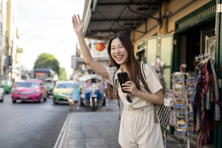 Téléchargez les photos : Jeune femme asiatique saluant un taxi dans la rue animée de Bangkok, Thaïlande Moment de voyage urbain authentique capturant la vie urbaine dynamique et la culture locale - en image libre de droit