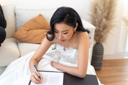 Photo for Asian woman in elegant bridal gown completing legal marriage paperwork. Bride in a white dress signs marriage documents, an official moment of commitment and beauty - Royalty Free Image