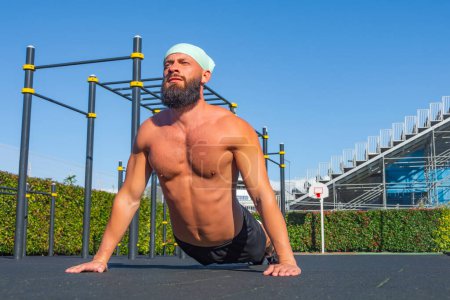 Téléchargez les photos : L'homme sportif fort pratique le yoga, fait des exercices d'étirement du dos et de renforcement du noyau le matin sur le terrain de sport par temps ensoleillé. Concept de santé et de fitness - en image libre de droit