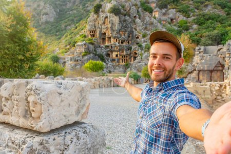 Photo for Swarthy caucasian european guy in plaid shirt smiles rejoices, takes selfie points with his hand show at the ancient unique landmarks Turkey, Antalya Demre, Myra, ancient lycian tomb casts faces - Royalty Free Image
