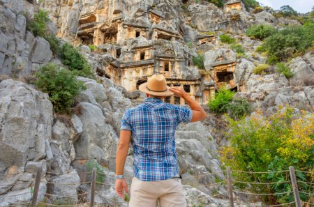 Photo for Swarthy caucasian european guy in plaid shirt and a hat stands back and looks in surprise at the ancient unique famous places Turkey, Antalya Demre, Myra, ancient lycian tomb casts faces - Royalty Free Image