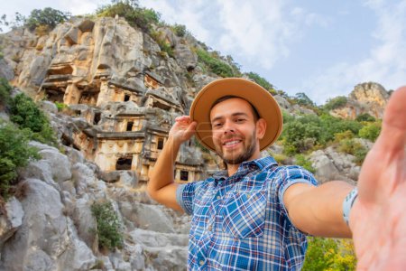 Photo for Swarthy caucasian european guy in plaid shirt smiles rejoices makes a selfie in a brown hat at the ancient unique landmarks Turkey, Antalya Demre, Myra, ancient lycian tomb cave ruins - Royalty Free Image