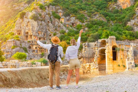 Photo for Guy with backpack in hat girl, travelers raising their hands up in surprise, look at ruins ancient city, ancient unique famous places of Turkey, Antalya Demre, Mira, ancient Lycian tomb casts faces - Royalty Free Image