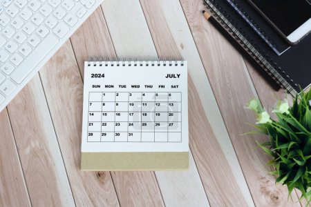 Photo for White July 2024 calendar on wooden desk with keyboard, note books, pencil, potted plant and smartphone. - Royalty Free Image