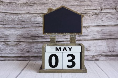 Chalkboard with May 03 calendar date on white cube block on wooden table.