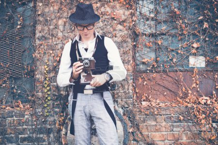 Foto de Fedora styled man using vintage analog photo camera while standing by brick wall. - Imagen libre de derechos