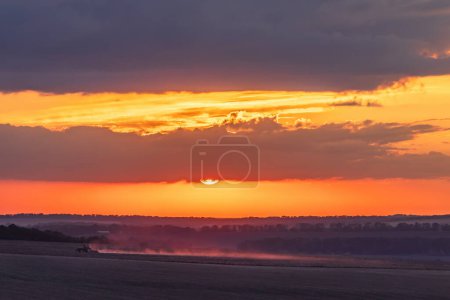 Foto de El tractor procesa los campos durante la puesta del sol y el polvo brilla en la oscuridad bajo el sol y el cielo colorido - Imagen libre de derechos