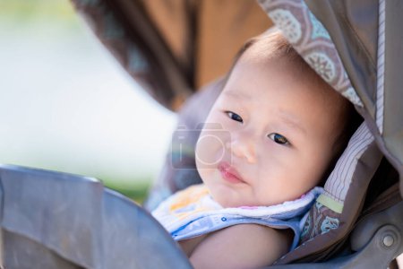 Téléchargez les photos : Bébé relaxant dans une poussette à l'extérieur. Enfant garçon avec un léger sourire se repose, regardant dehors avec des yeux curieux, profiter du confort et des vues d'une journée tranquille à l'extérieur. Asiatique Enfant de 1 an. - en image libre de droit