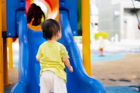 Téléchargez les photos : Retour de bébé garçon asiatique avec toboggan coloré et swing à l'aire de jeux. Un enfant d'un an se tient à la fin du toboggan bleu. Les enfants jouent à l'école ou à la maternelle. Activité estivale saine pour les enfants. - en image libre de droit