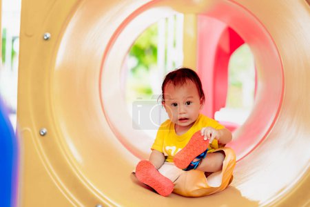 Portrait of a one-year-old boy taking off his shoes while sitting in a toy tunnel in playground, Child play and climb for fun, Toddler sweat from the hot weather, Baby is growing up strong,Summer time