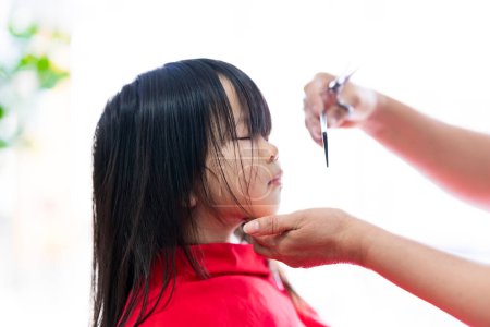 The hands of a barber meticulously cut the hair of a cute Asian girl.