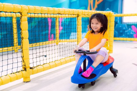 Adorable fille asiatique chevauchant voiture jouet bleu dans une aire de jeux intérieure, souriant et s'amusant. Enfant de 7 ans.